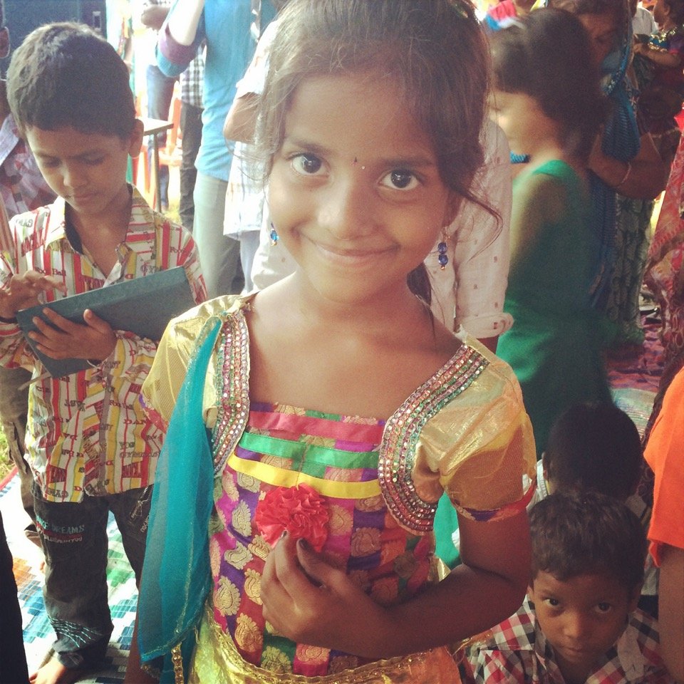 A smiling young girl in colorful traditional dress at a crowded event, holding a red flower, with children and adults in the background.