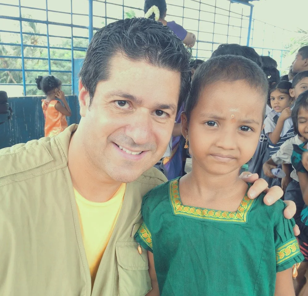 A man smiling and taking a selfie with a young girl who has a serious expression, in a classroom or community space with children in the background.