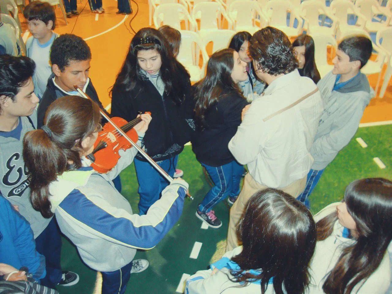 A group of students gathered in a circle listening to a woman speak, with one student playing a violin. The setting appears to be a gymnasium.