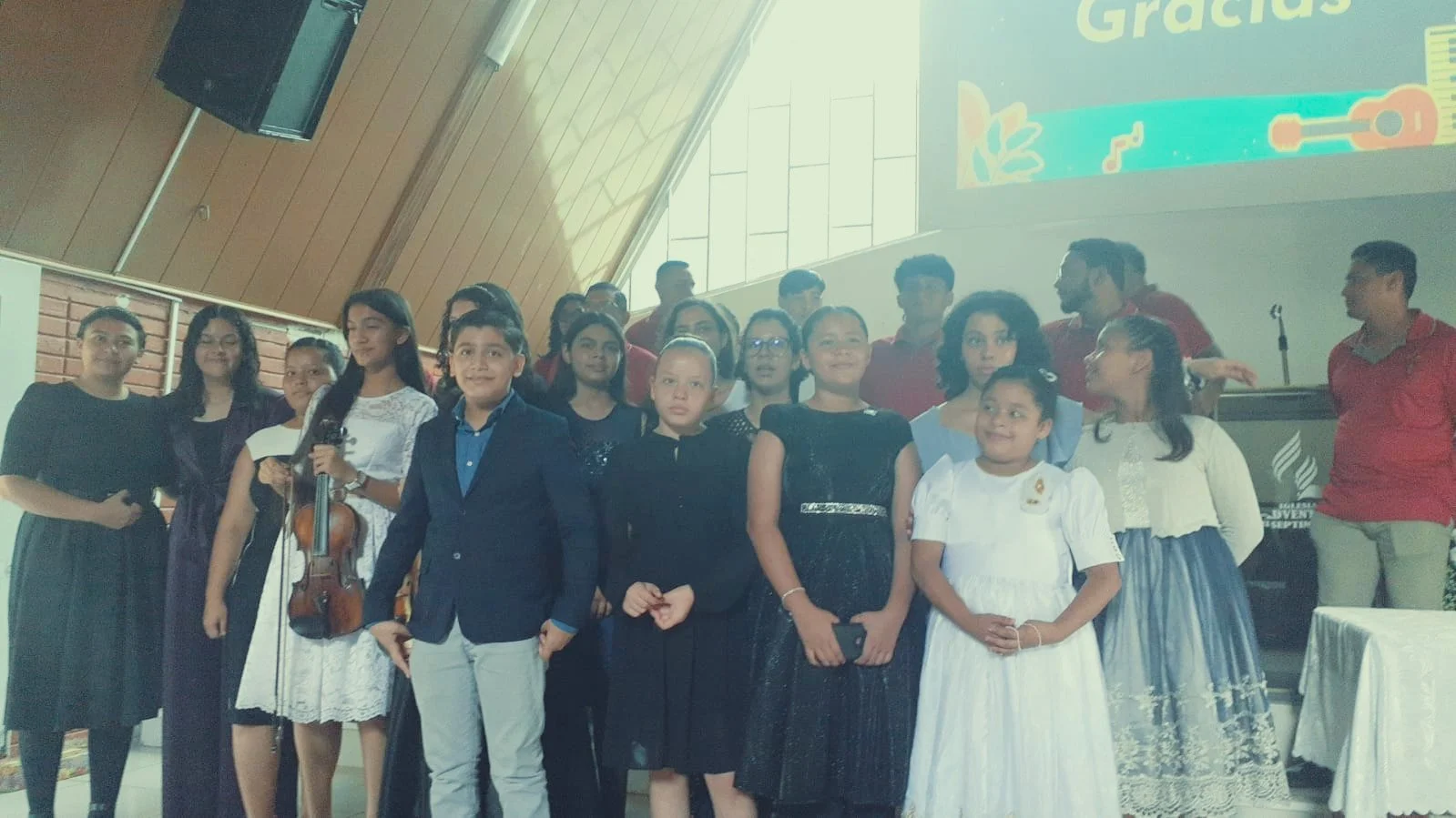 Group of children and young adults on stage during a graduation or school event, some wearing formal attire, with a large screen in the background.