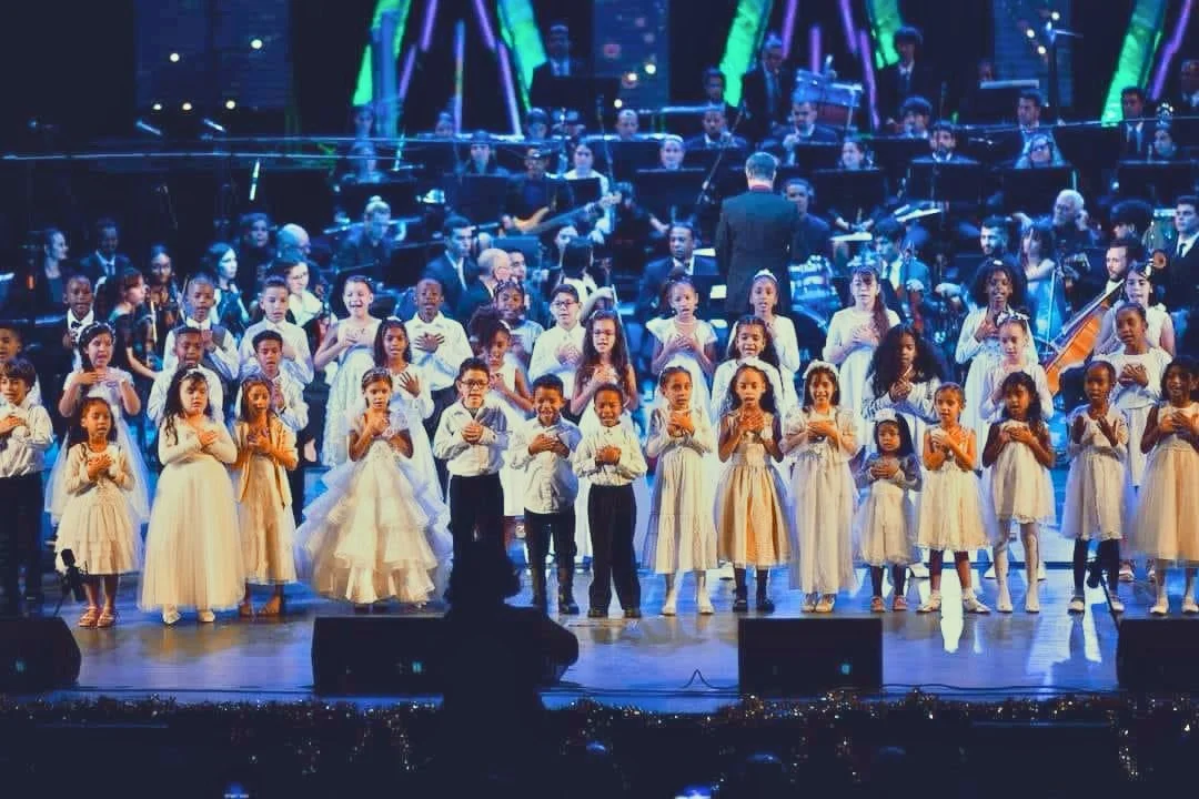Children in white dresses and shirts singing on stage during a concert, with an orchestra in the background.