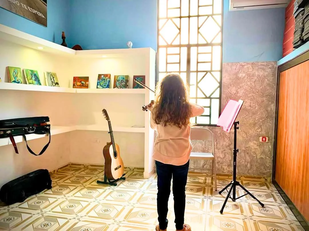 A girl with curly hair playing the violin in a room with musical instruments and artwork, sunlight through a geometric window casting bright light.