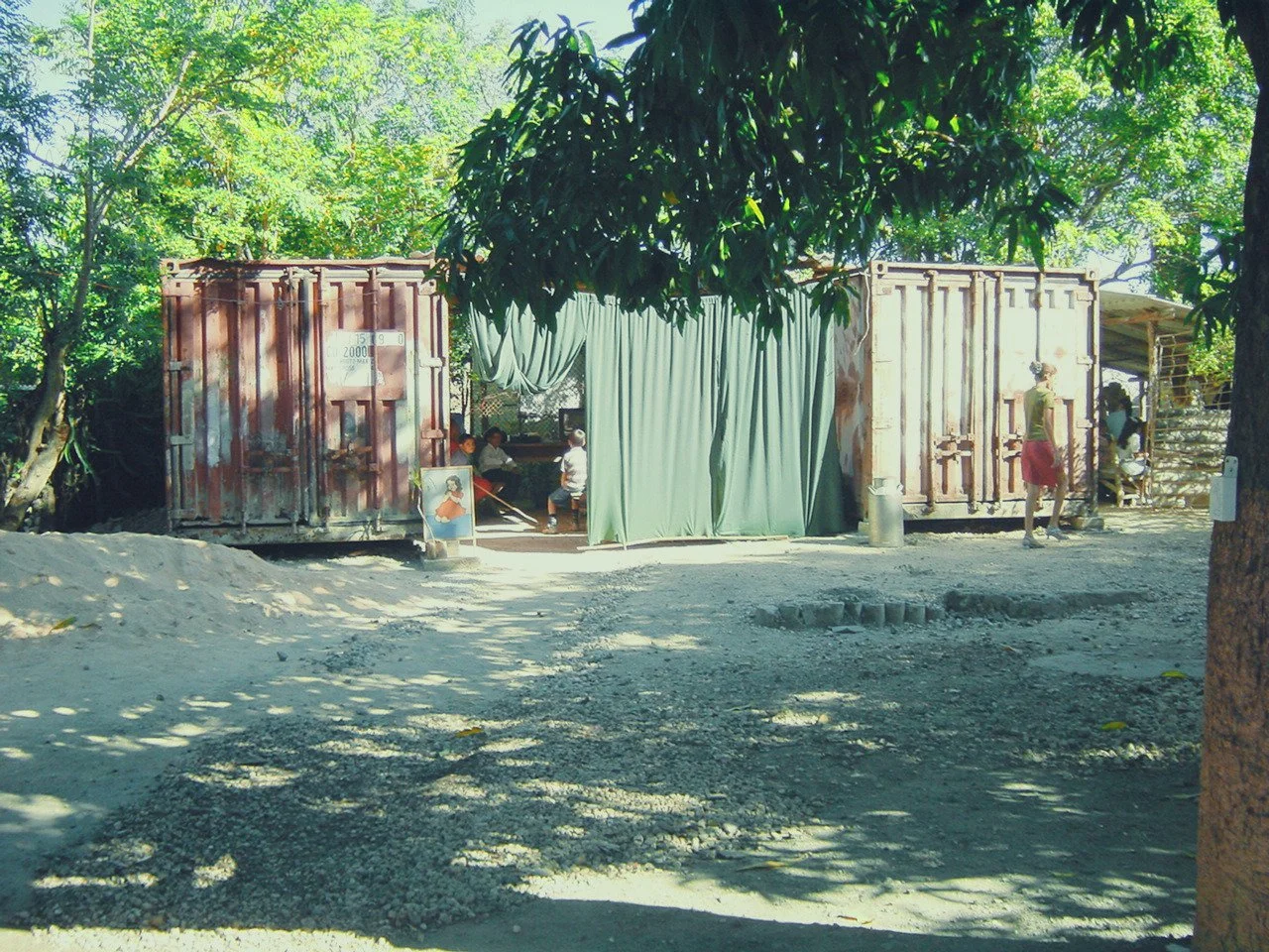 A makeshift outdoor seating area with a green curtain at the entrance, flanked by two shipping containers. There are trees providing shade, and some people are sitting and standing around the area.