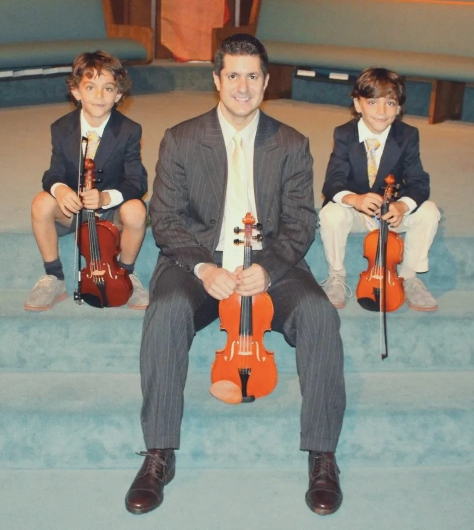 A man in a suit sitting with two young boys in formal attire, all holding violins and smiling, posed on a carpeted steps in an indoor setting.