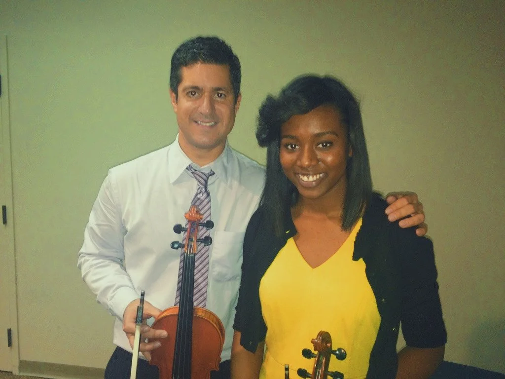 A man holds a violin and a bow, standing next to a woman in a yellow top and black jacket. Both are smiling and standing indoors against a neutral background.