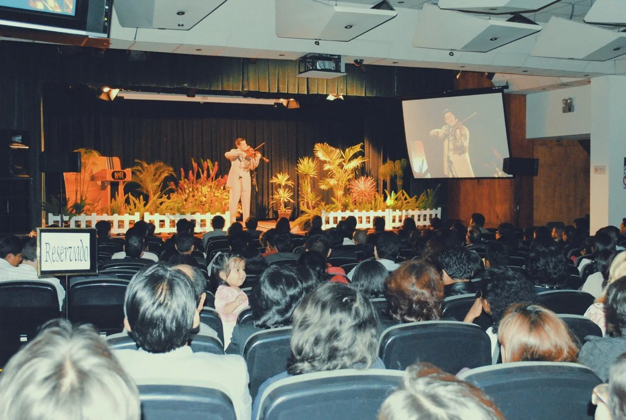 An indoor auditorium with a stage decorated with plants and a white fence, where a person is performing a violin. A large screen shows a close-up of the performer. Audience members are seated, watching the performance, and a reserved sign is visible 
