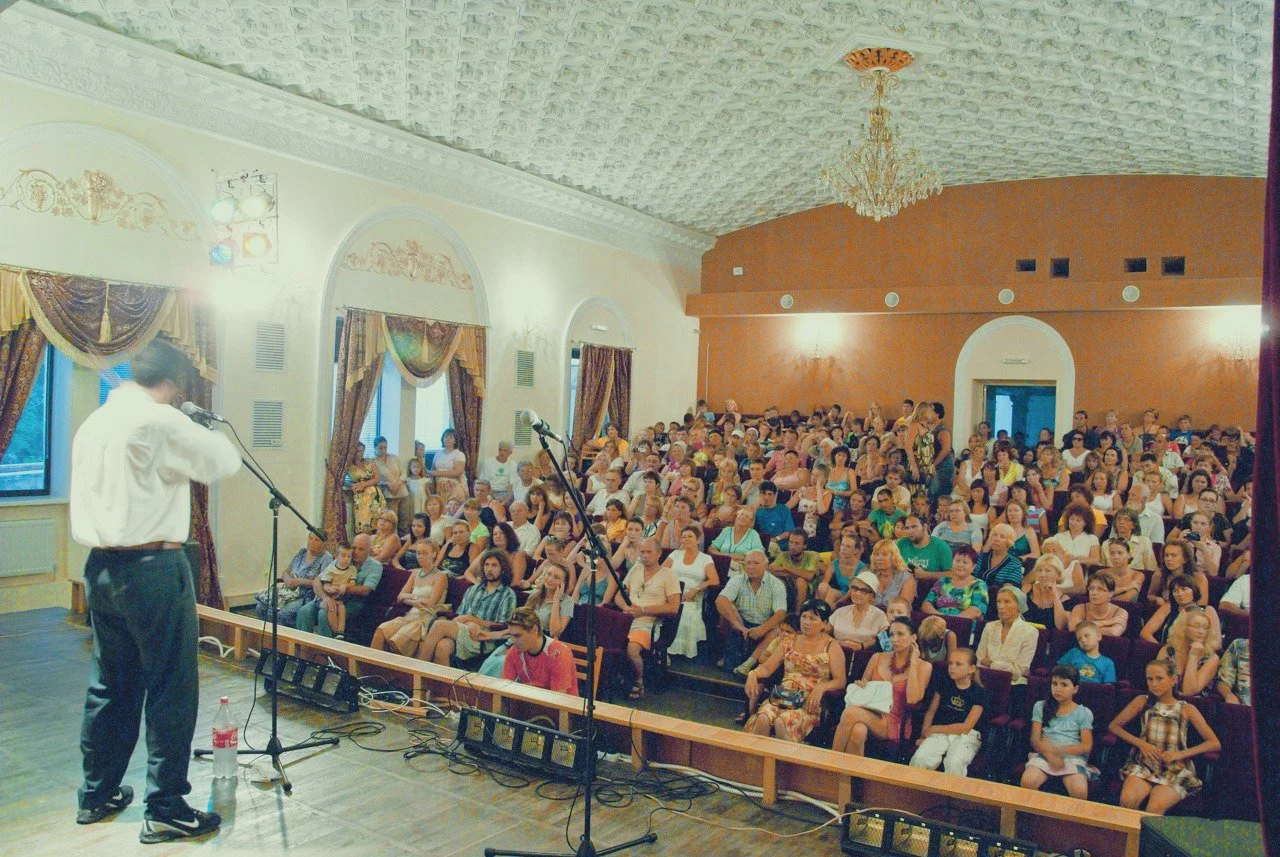 A man on stage speaking into a microphone in front of a large audience seated in an ornate hall with chandeliers and decorative curtains.