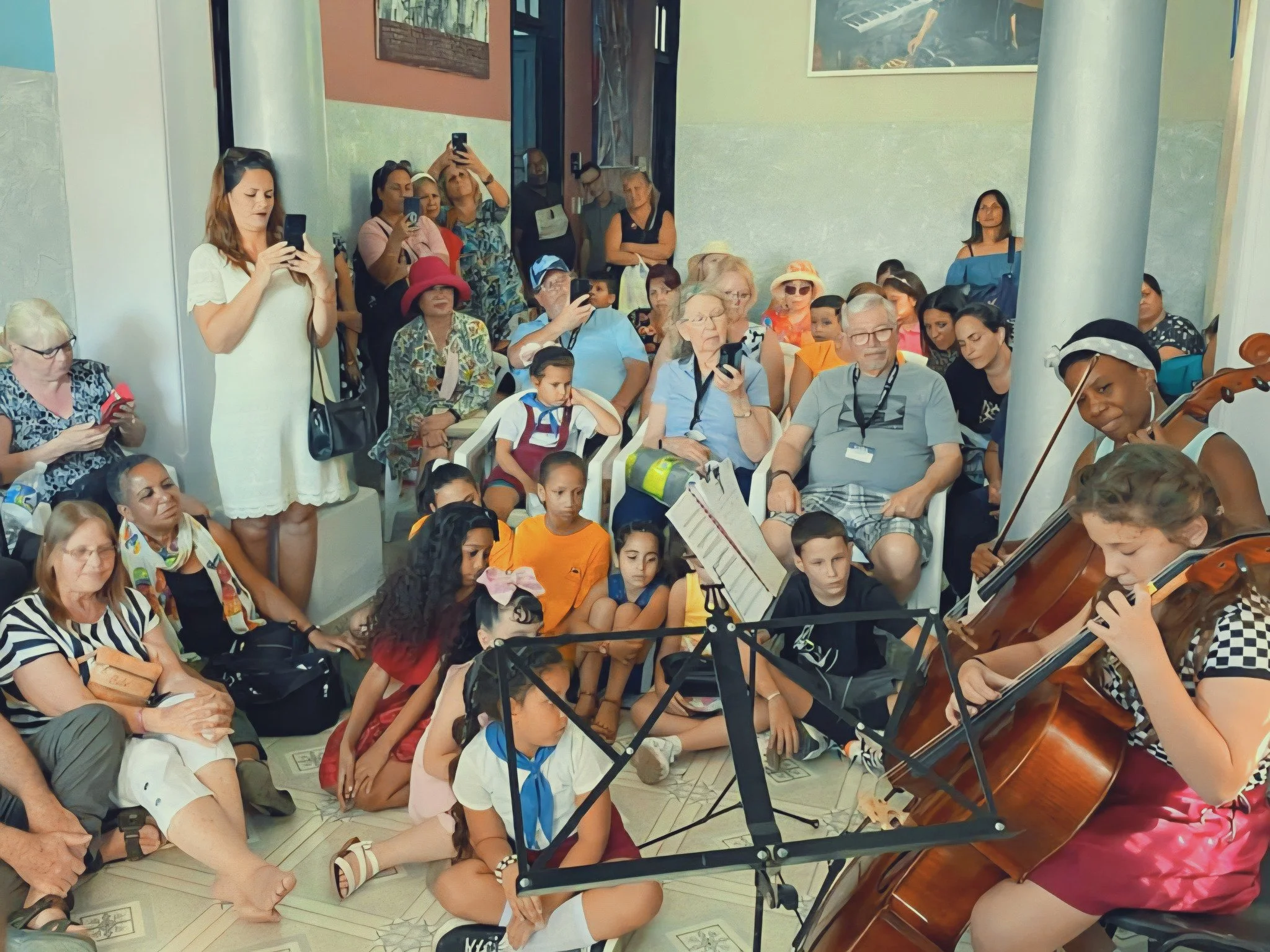 A large group of people, including children and adults, gathered indoors with some seated on chairs and some standing, as a woman plays the violin in the foreground. Several people are taking photos or videos with their phones, and the setting appears to be an event or gathering.