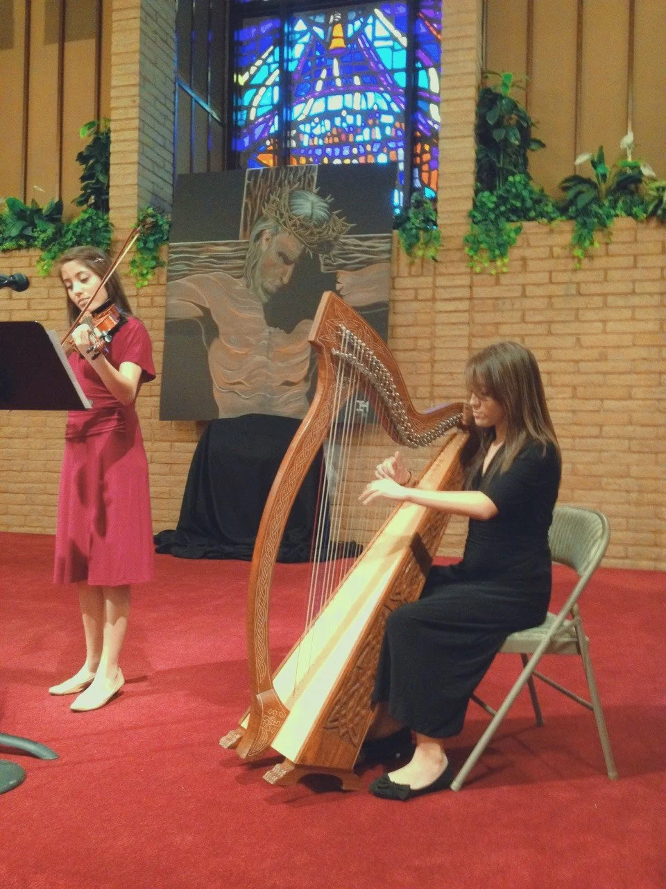 Two women performing music inside a church with stained glass windows and a painting of Jesus Christ in the background. One woman is playing a violin, and the other is playing a harp.