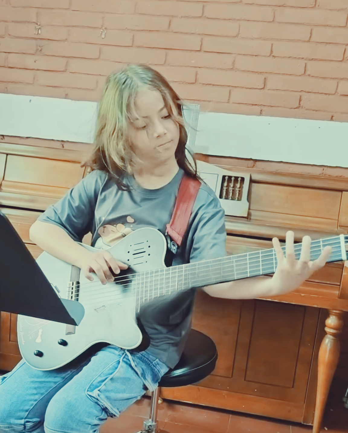 A young girl with multicolored hair plays an acoustic guitar while sitting on a stool in a room with a brick wall background.