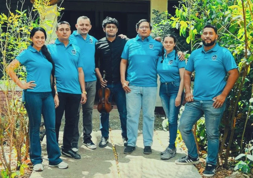 Group of seven people standing outdoors, some holding a violin, all wearing matching blue shirts, in a garden setting with plants around.