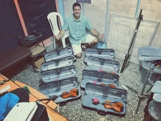 A man sitting on a white chair in a room with a blackboard, surrounded by multiple open violin cases with violins inside, and some tools or accessories.