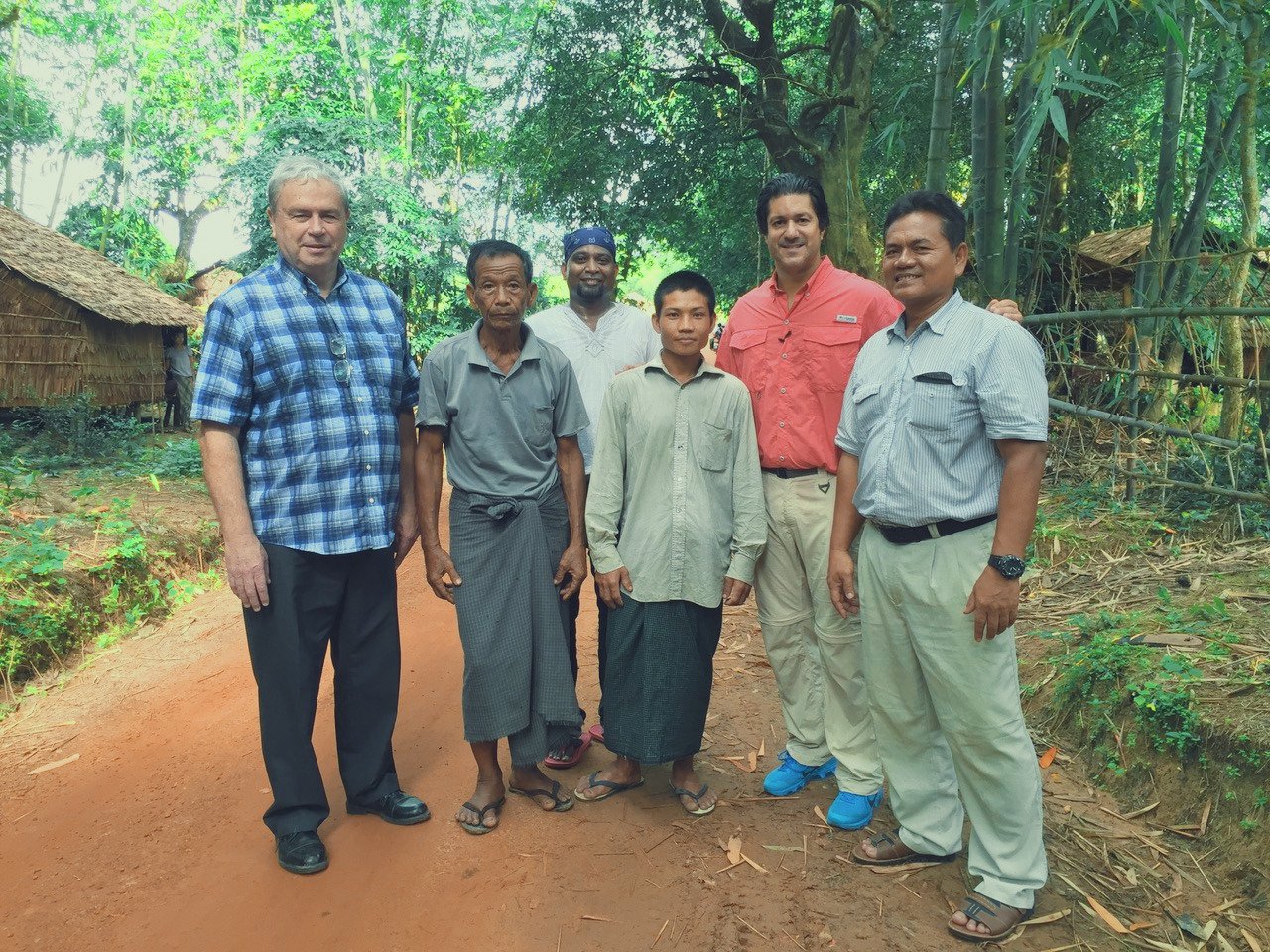 Six men standing on a dirt path in a lush green jungle, with bamboo and traditional huts in the background.