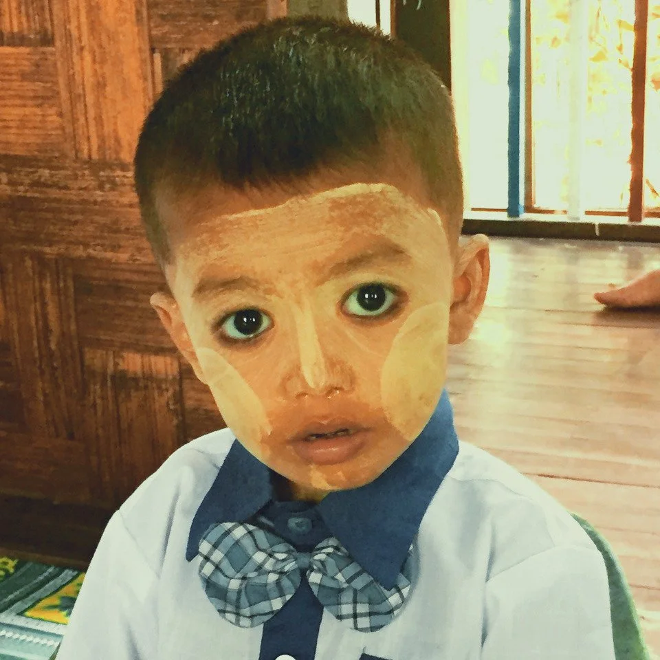 A young boy with traditional tan face paint and large, expressive eyes. He is wearing a white shirt with a blue collar and a plaid bow tie, sitting indoors with wooden walls and a window in the background.