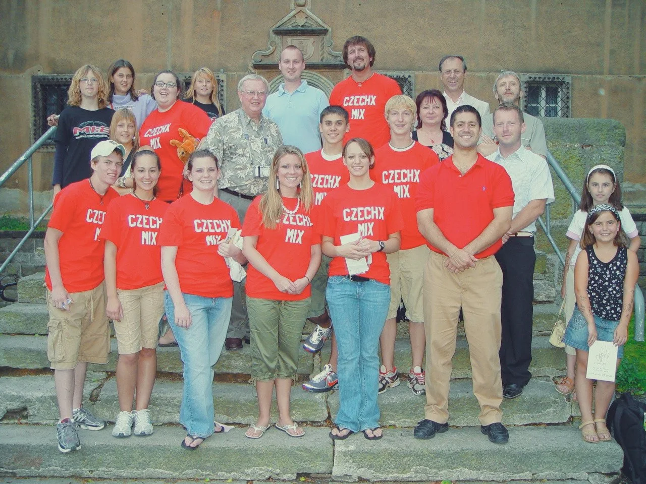 Group photo of people on steps, some wearing red shirts with 'CZECHX MIX' printed on them, in front of a historic building with stone walls and windows.