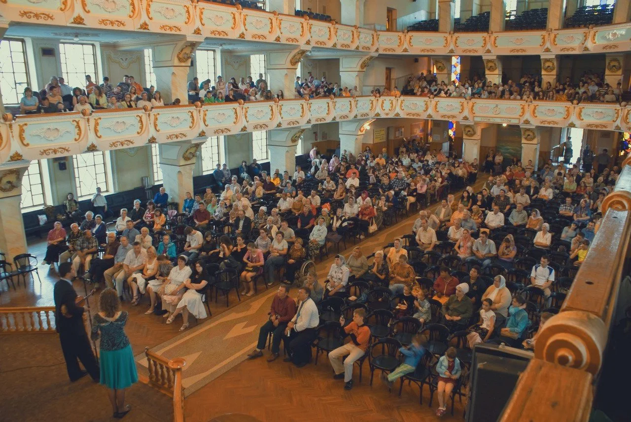 Large audience seated in a decorated theater or hall, watching a performance or presentation, with two performers on stage.