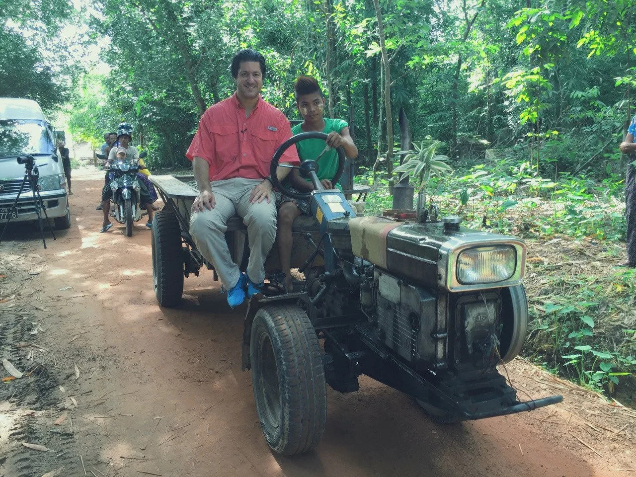 Two people sitting on a makeshift tractor in a forested area, with a man in a red shirt and a young girl in a green shirt holding the steering wheel, surrounded by a dirt path and some parked vehicles and motorcycles in the background.