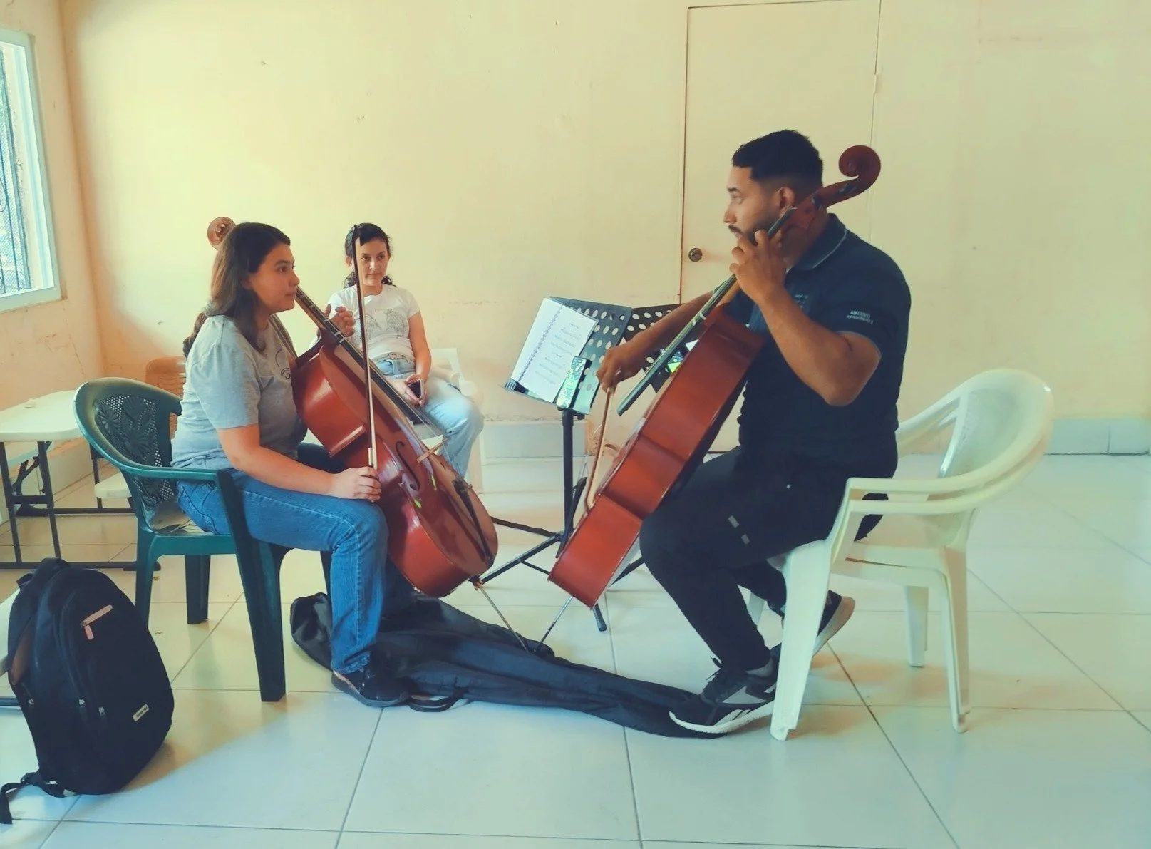 Two young girls and one adult man practicing cello playing in a room, with music stands and sheet music.