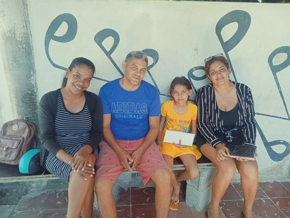 Four people sitting on a bench against a painted wall with musical notes, including two women, a man, and a young girl holding a small board.