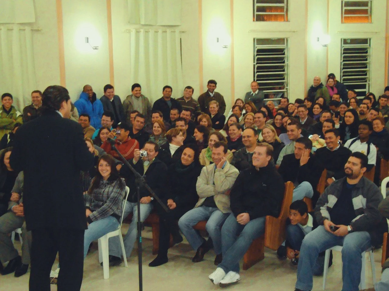 A man in a black suit speaks to a large, seated audience in a hall with vertical orange and cream-colored walls and multiple windows.