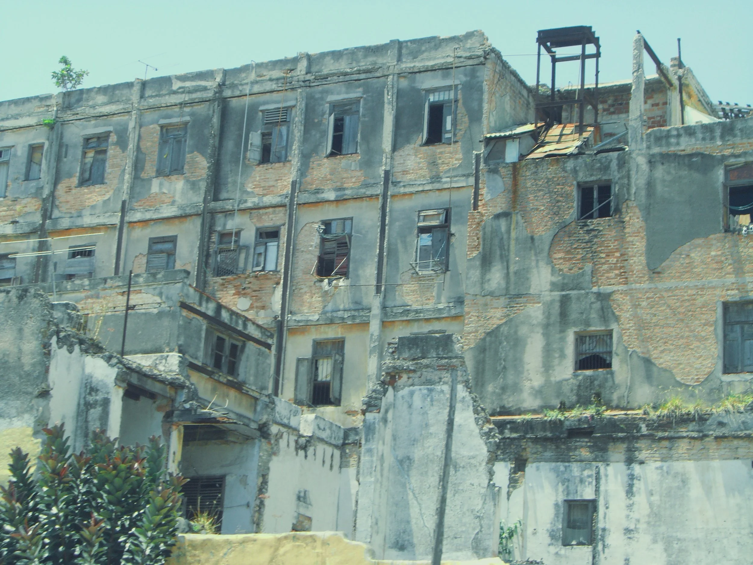 An old multi-story building with peeling paint, exposed brick, broken and missing windows, some with open shutters, and a metal staircase on the side.