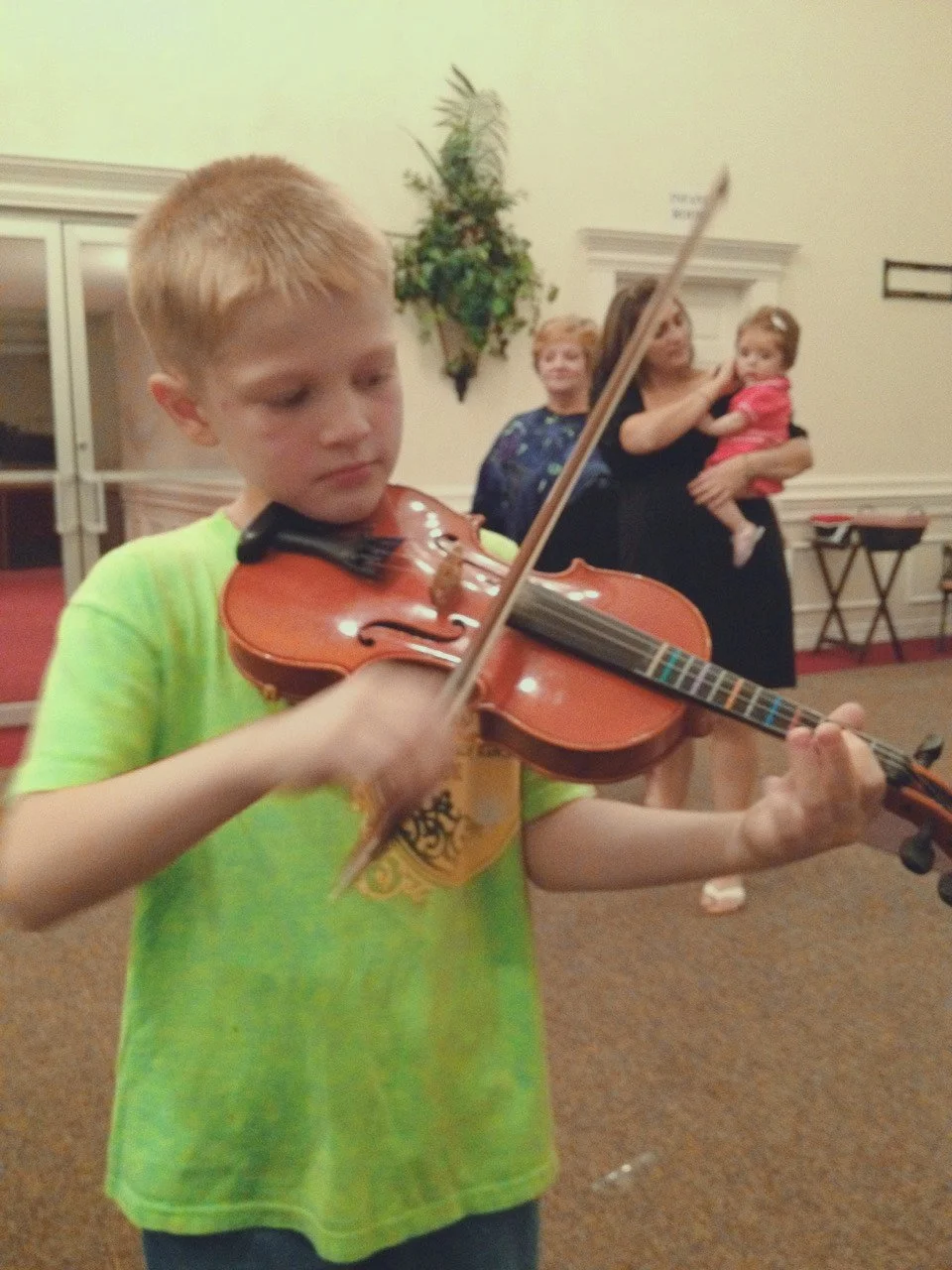 Young boy playing a violin with adults in the background in an indoor setting.