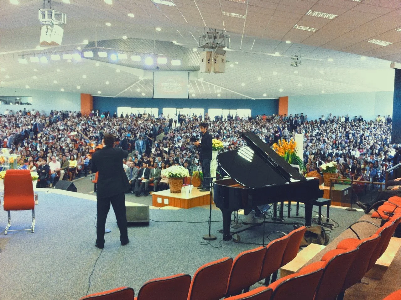 A large indoor church service with many people seated, a man speaking at a podium, a person playing piano, and floral decorations on stage.