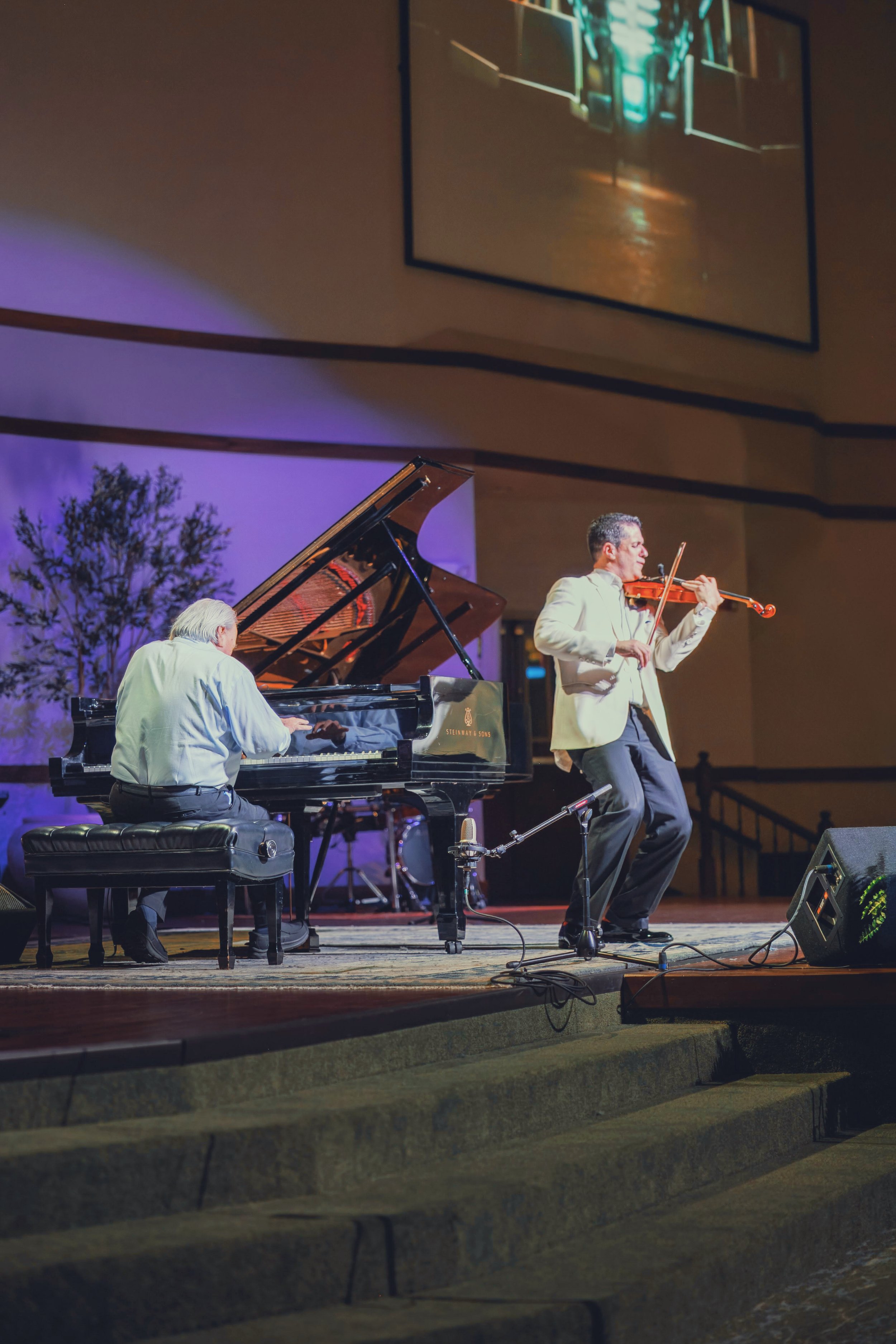 Two musicians performing on a stage: a man playing the piano and a man playing the violin, with a large screen showing their performance in the background.