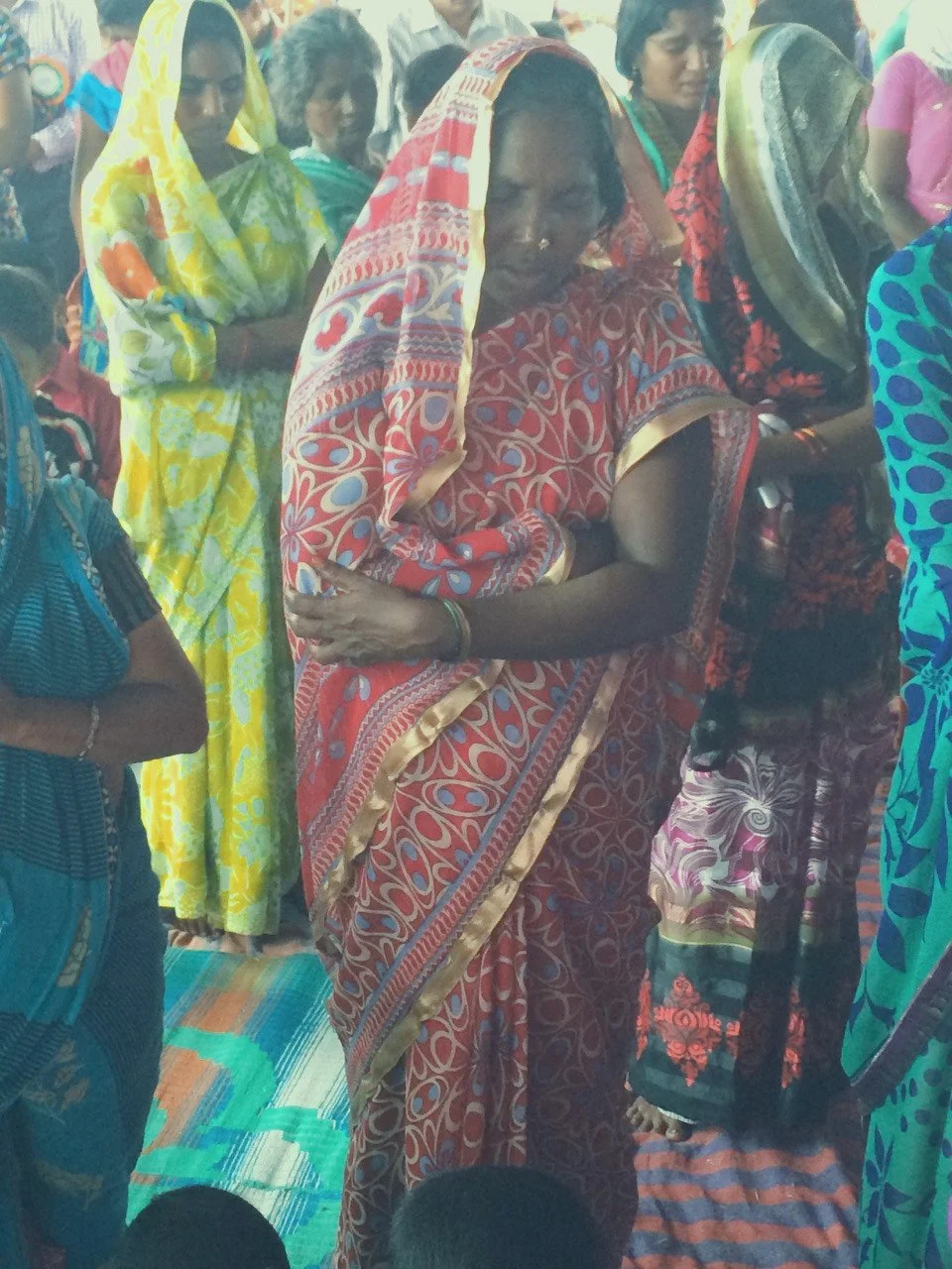 A group of women, mostly wearing colorful traditional sarees, standing close together indoors, with some women looking downward and others looking ahead. The central woman is dressed in a red patterned saree with gold trim, with a nose ring, in a cro