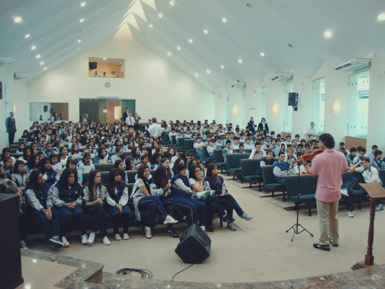 A young musician playing the violin on stage in front of a large audience, mostly school students, in a spacious auditorium.