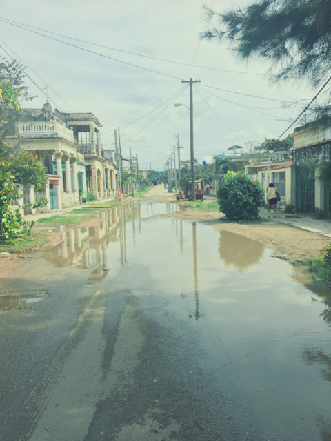A flooded street with water covering the road, surrounded by residential buildings, trees, and utility poles.