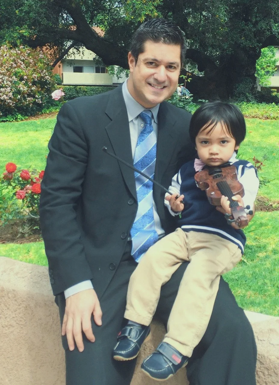 A man in a suit and a young boy sitting on a stone, holding a toy violin and bow, outdoors in a garden with flowers and trees.
