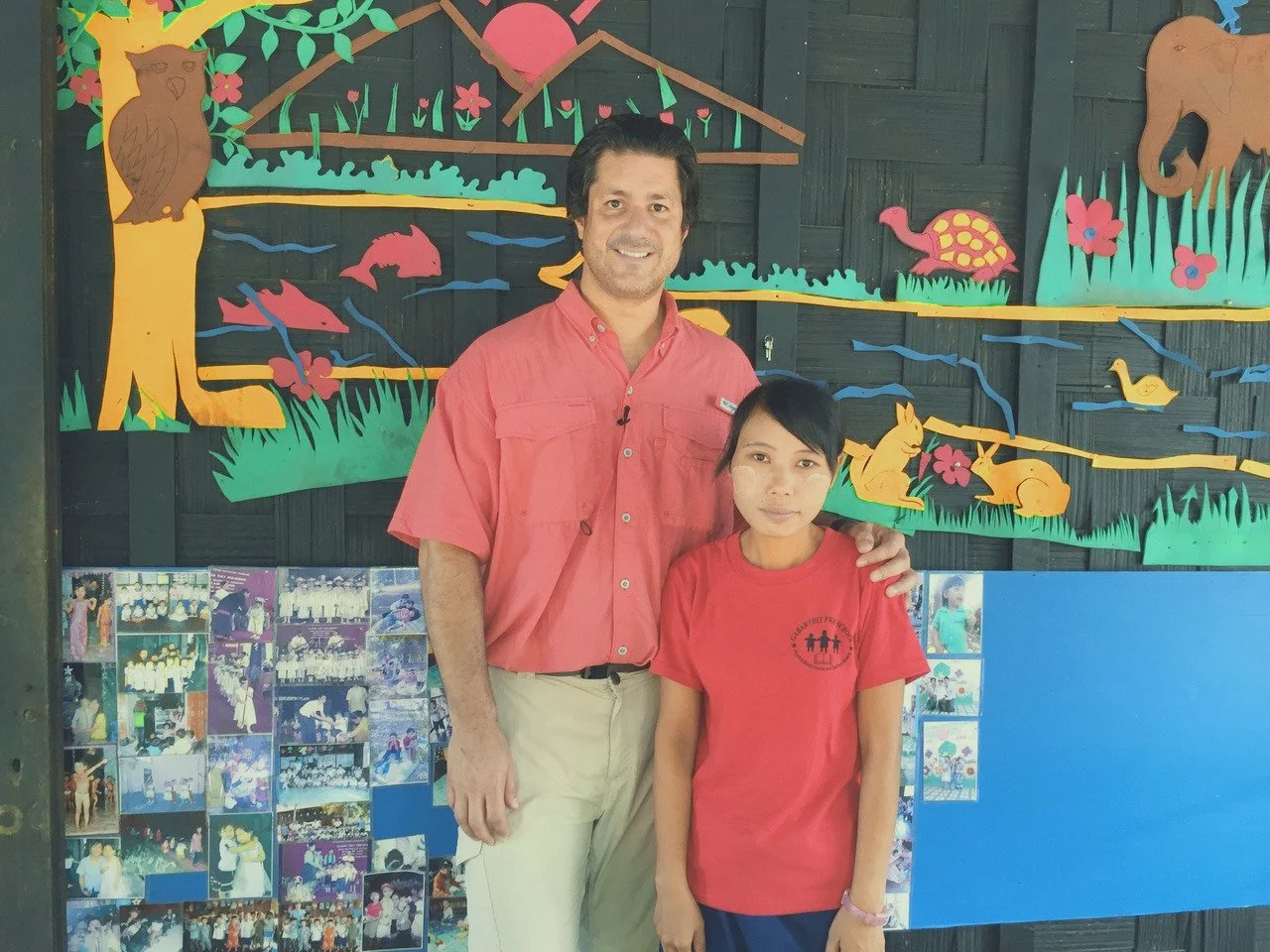 A man and a young girl standing in front of a colorful paper cutout mural of a nature scene with animals, trees, and a pond, with a collage of photographs below.