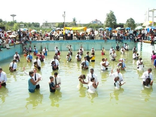A large crowd gathered around a pool filled with water, where many people are standing in the water and participating in a religious or cultural ritual, with some holding objects in prayer. Spectators with umbrellas line the edges and background, and