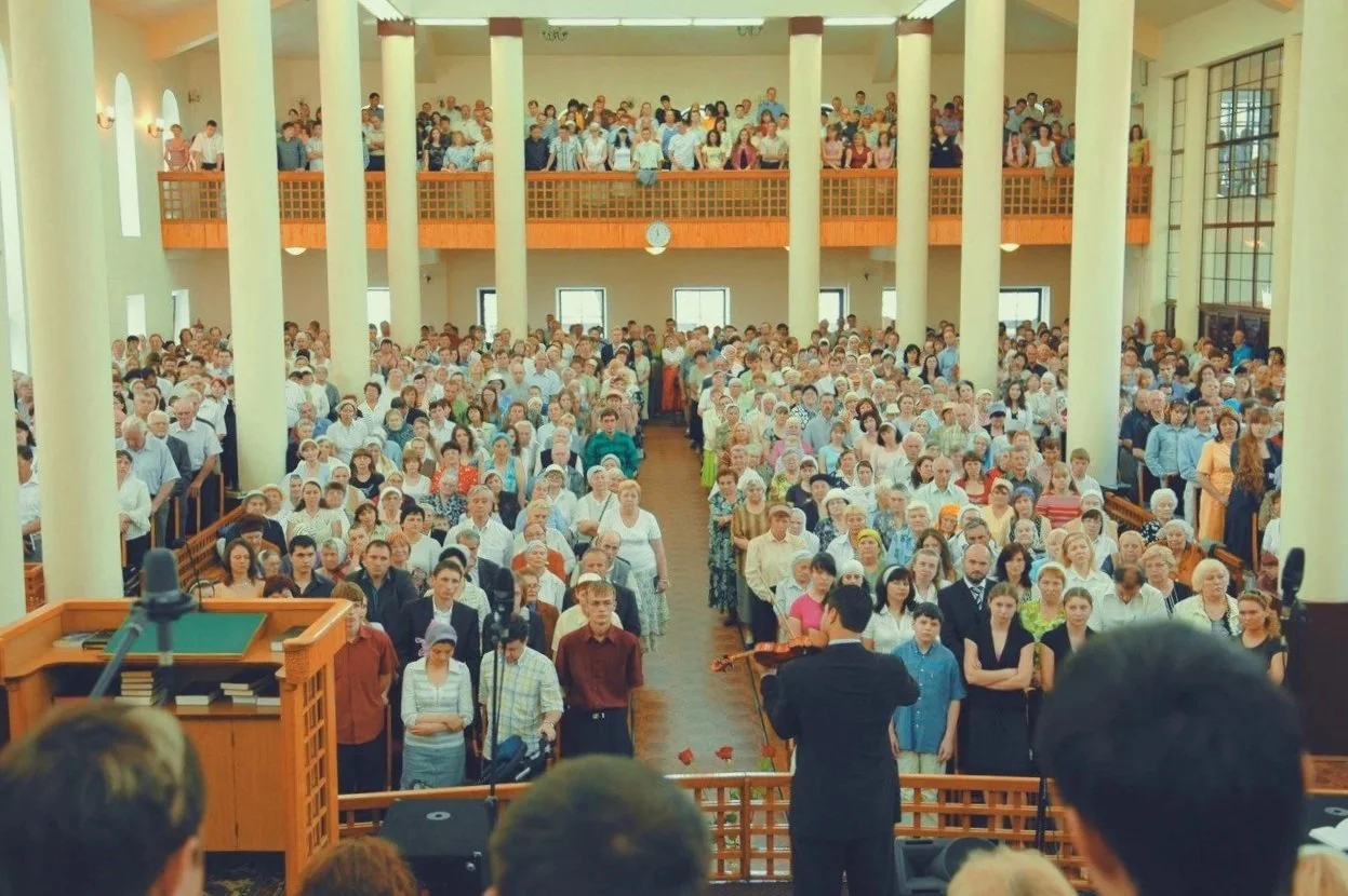 A large congregation gathered in a spacious church with high ceilings, white columns, and an upper balcony. People are standing and facing the front, where a person is playing a violin.