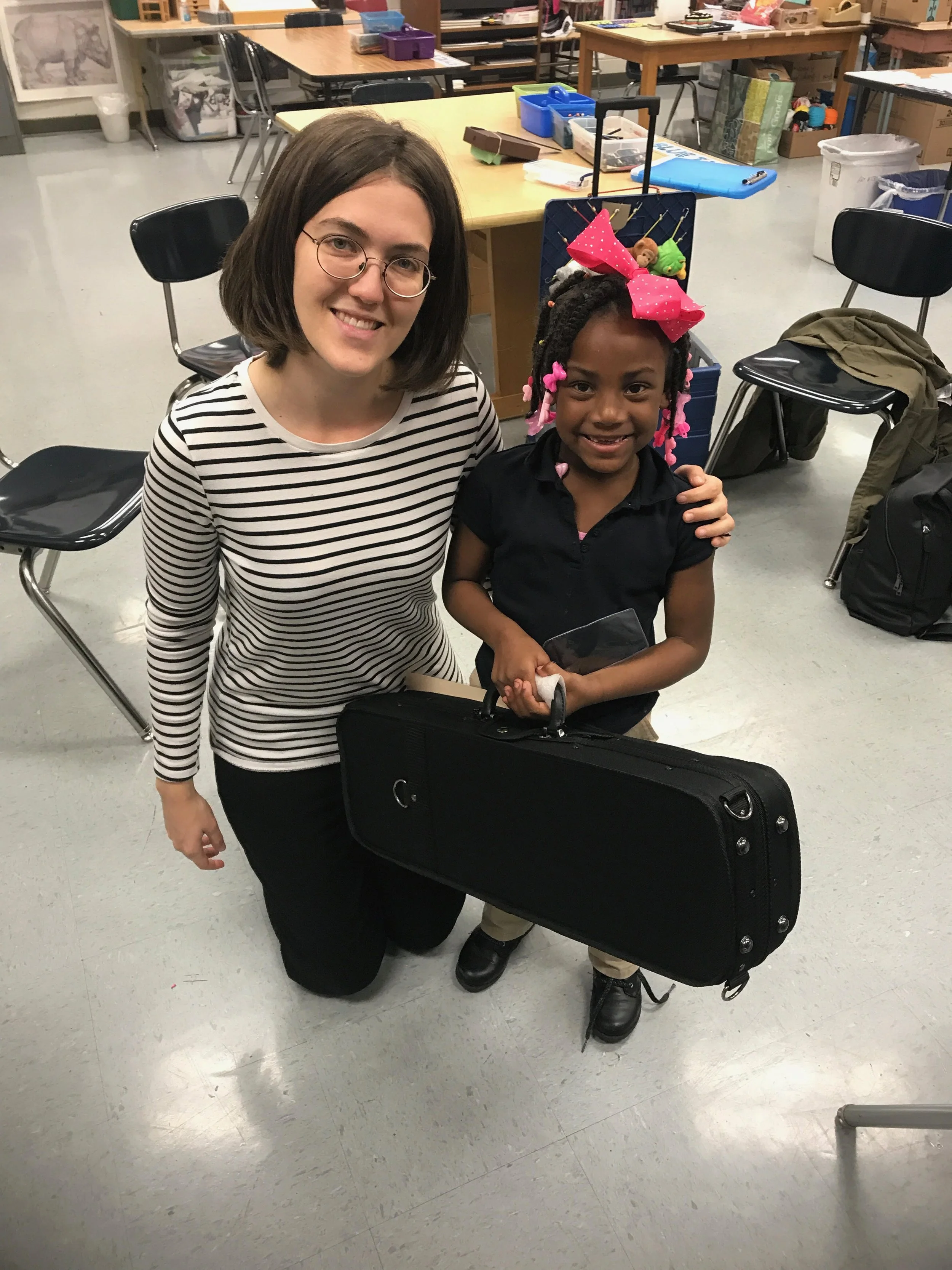 A woman kneeling on the floor, smiling, next to a young girl with a pink bow in her hair, standing indoors in a classroom or office setting with tables, chairs, and various supplies in the background.