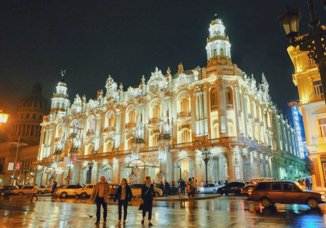 Historic building illuminated at night with people walking on wet street in front and cars parked nearby.