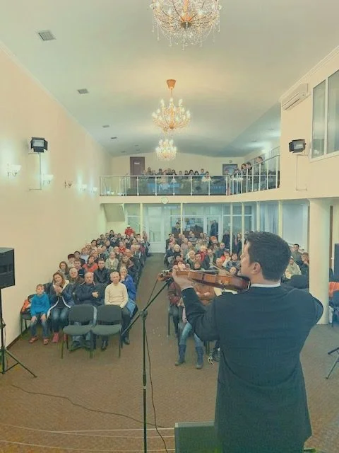Violinist performing in front of a seated audience in a spacious room with chandeliers and a second-level balcony.