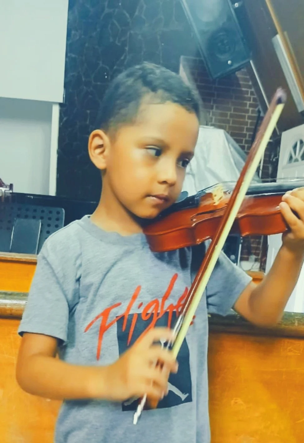 A young boy playing a violin indoors, standing in front of a brick wall and wooden counter, focused on his instrument.