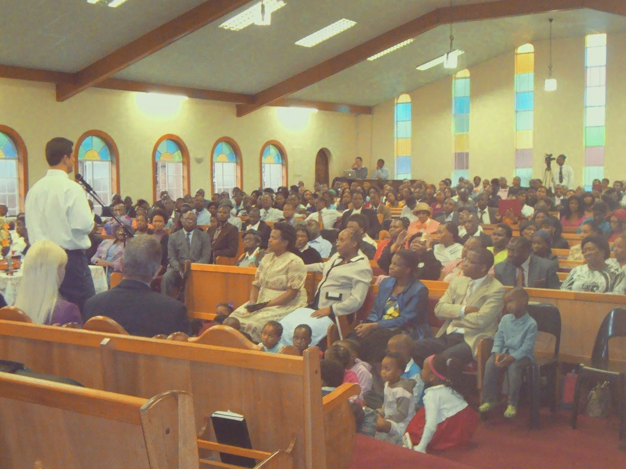 A large congregation gathered inside a church with stained glass windows, where a man is speaking at the front.