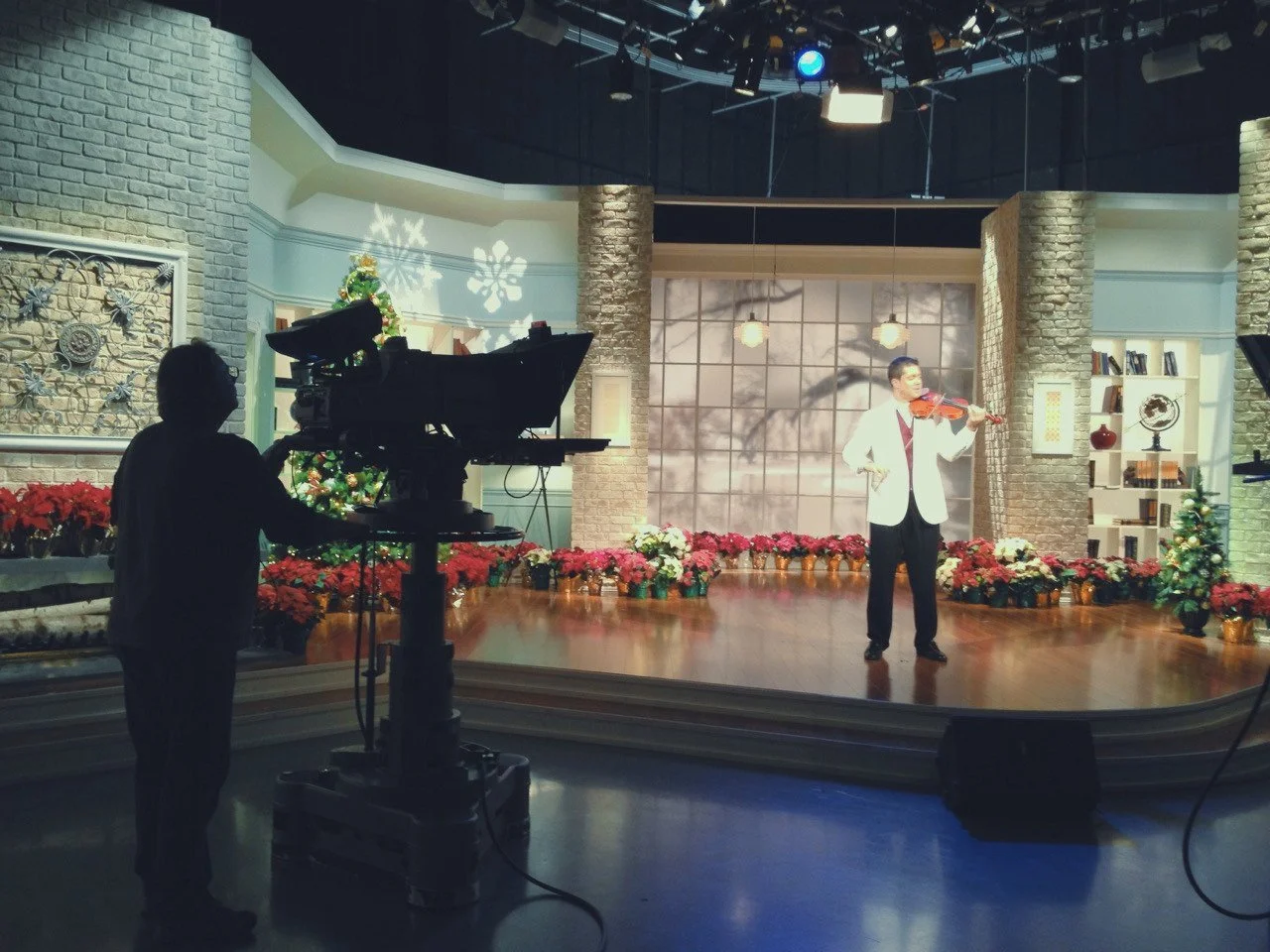 TV studio set with a man playing a violin on a decorated stage, camera operator filming, holiday decorations, Christmas tree, and floral arrangements