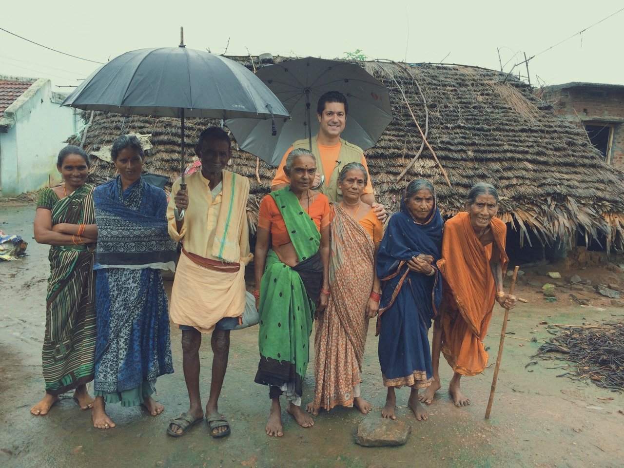 A group of nine people, including women and men, standing in front of a thatched roof hut with mud walls, some holding umbrellas, in a rural setting. They are dressed in traditional Indian clothing, and some are barefoot, with a muddy ground.