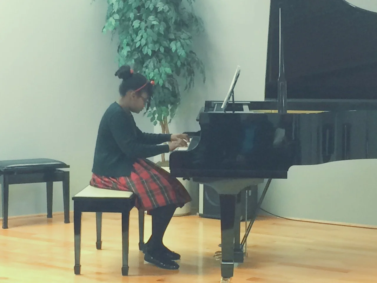 A young girl with glasses and a bun hairstyle, wearing a black sweater and a red plaid skirt, sitting on a padded bench, playing a grand piano in a room with hardwood floor and a tall potted plant in the background.