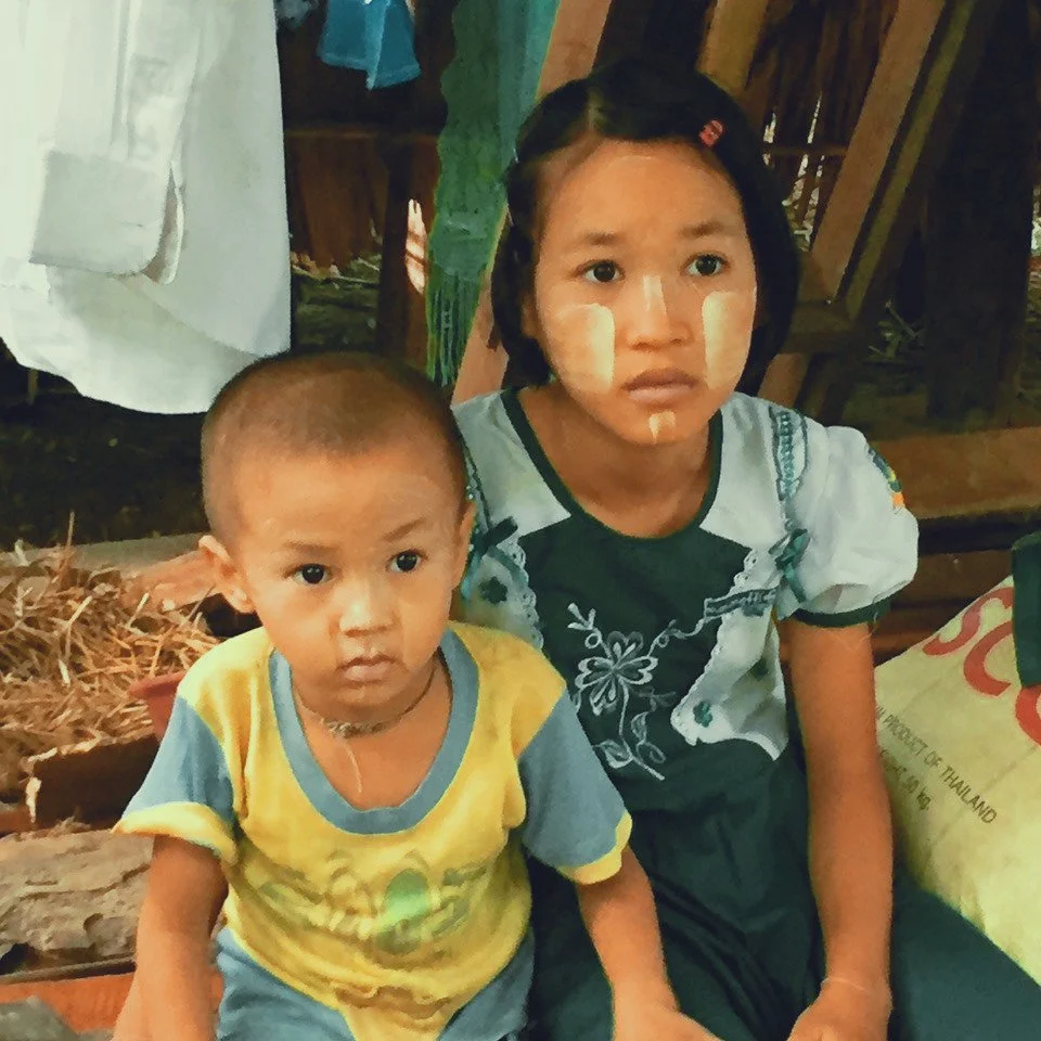 Two children, a girl and a boy, sitting indoors with wooden walls and a sack to the right. The girl has traditional white face paint and is wearing a light blue dress with dark blue accents. The boy has a shaved head and is wearing a yellow and blue 