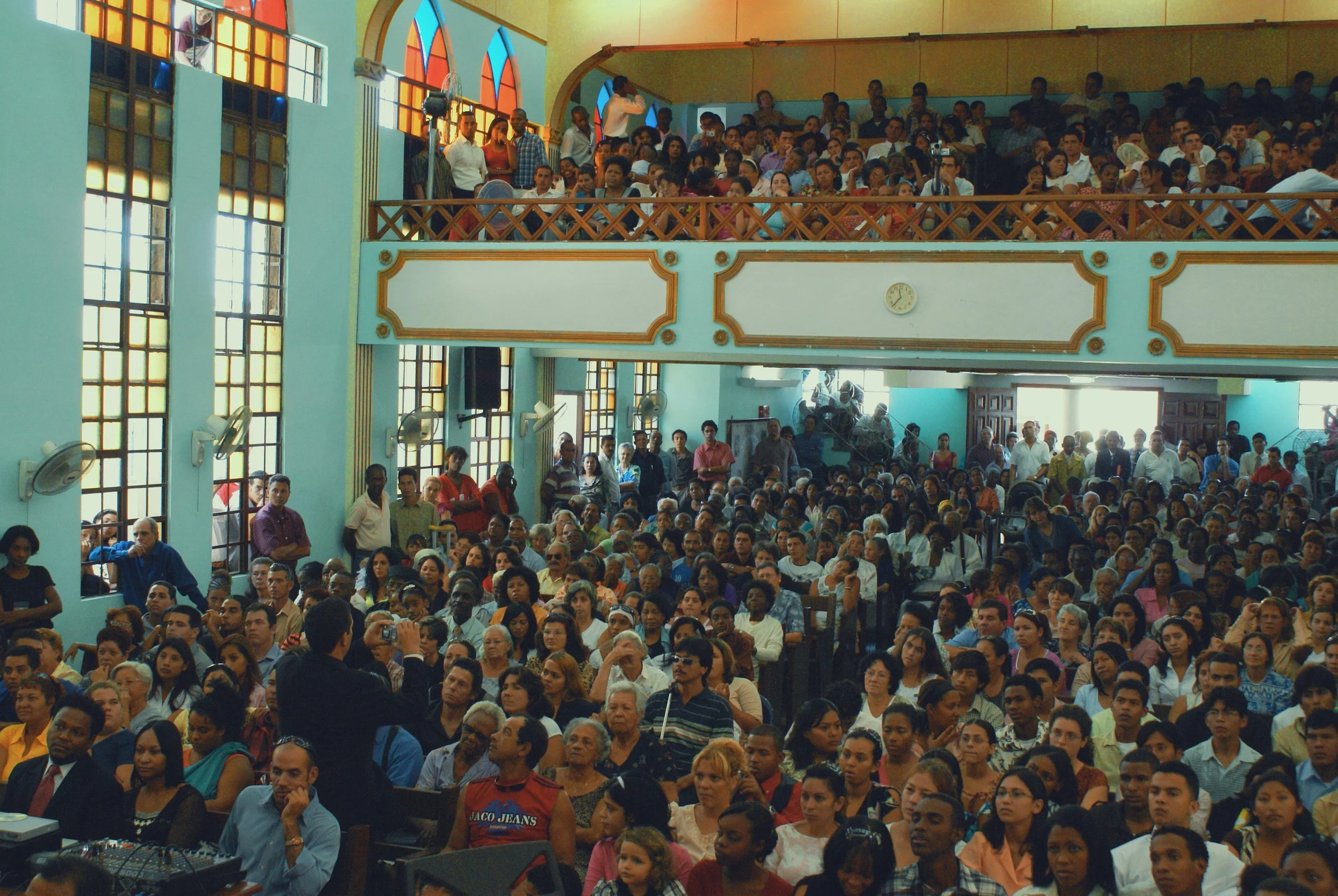 A large indoor gathering with many people seated and standing, inside a hall with tall stained glass windows, a clock on the wall, and arched architectural features.