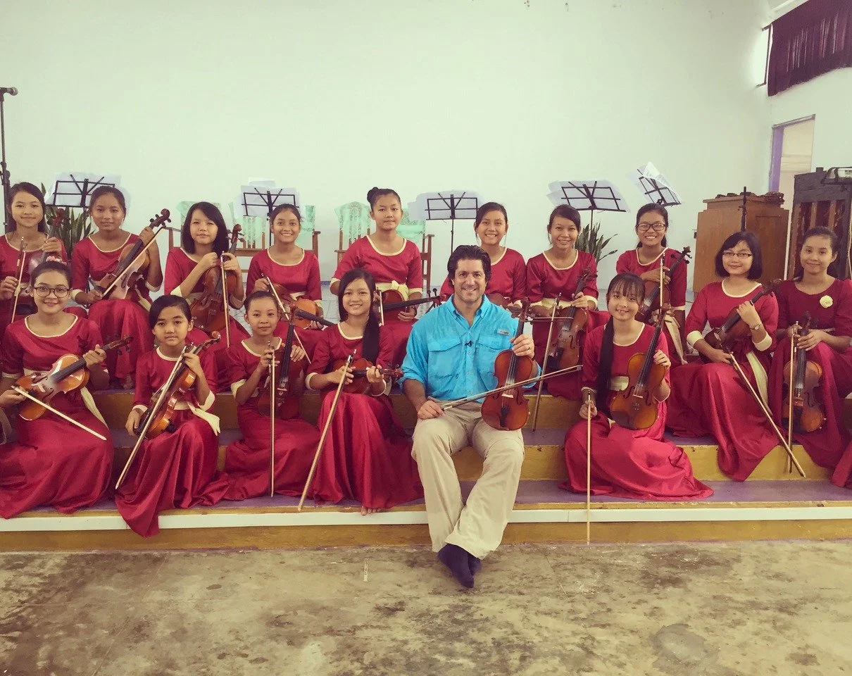 A group of young girls in red dresses holding violins and bows, seated on a stage with sheet music stands behind them, and a man in a blue shirt sitting in front, holding a violin and bow, in a music classroom or rehearsal space.