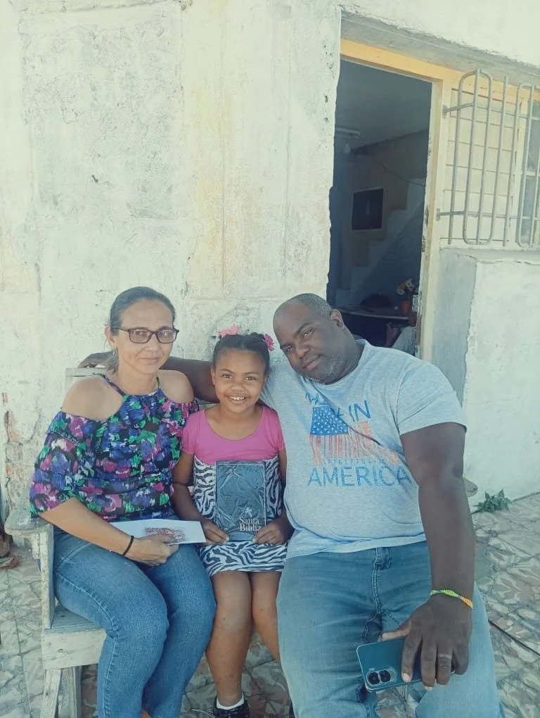 Three people sitting outdoors in front of a white wall, a woman and a man with a young girl in between, holding a Bible, smiling.