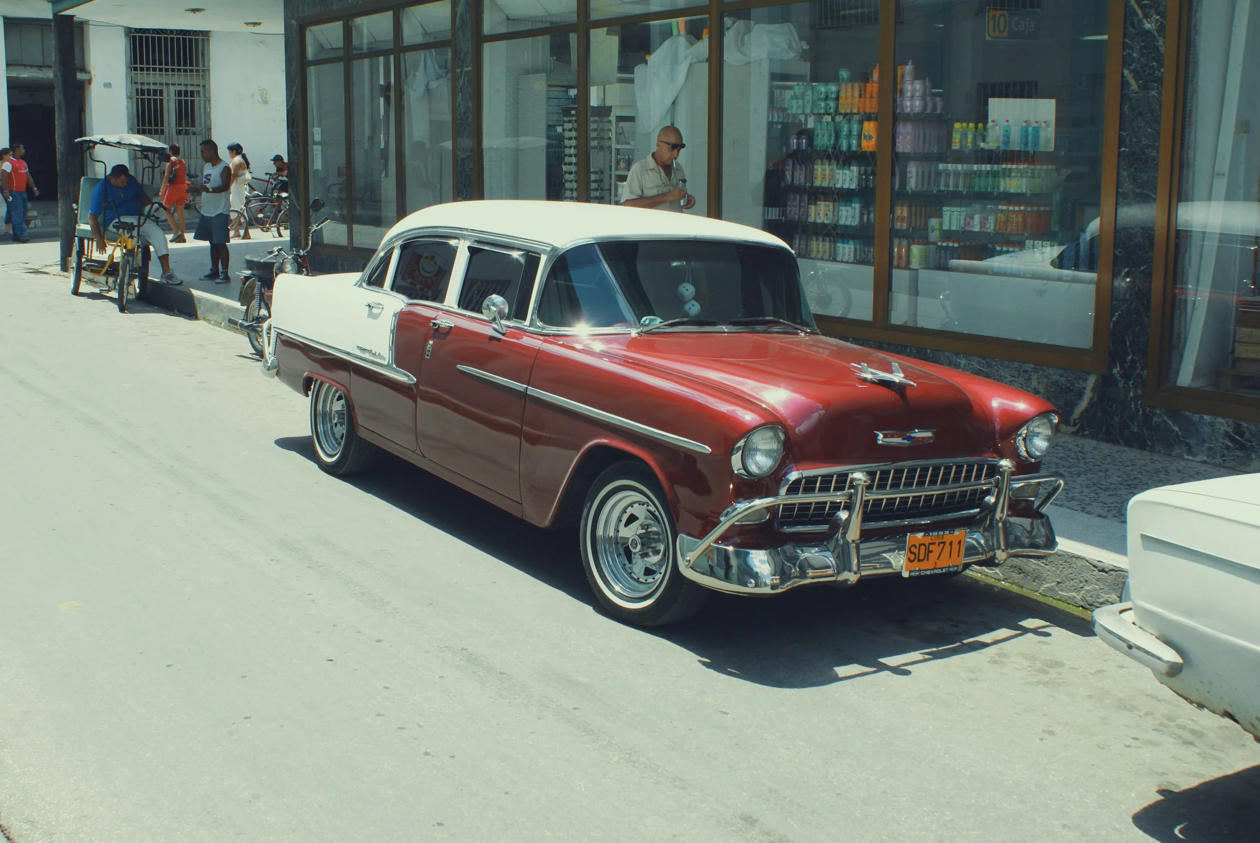 A vintage red and white Chevrolet car parked on the street in front of a store with glass windows. There are people walking and riding bicycles nearby, and a man inside the store reading a newspaper.