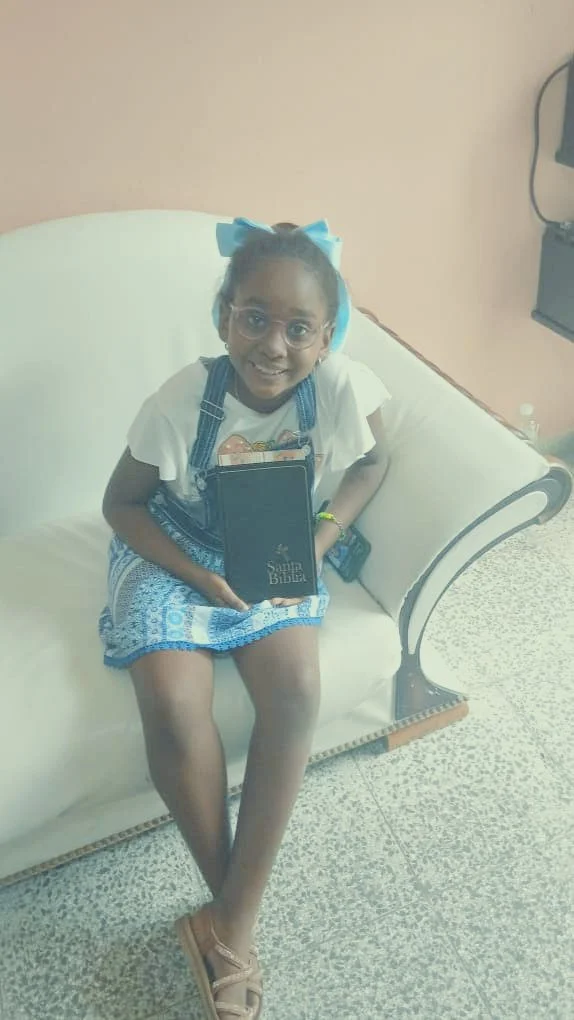 A young girl sitting on a white cushioned vintage-style sofa holding a black book titled "Sabe Bibbia" with a big smile, wearing glasses, a white t-shirt, a blue patterned skirt, and a blue bow in her hair, in a room with cream-colored walls and patt
