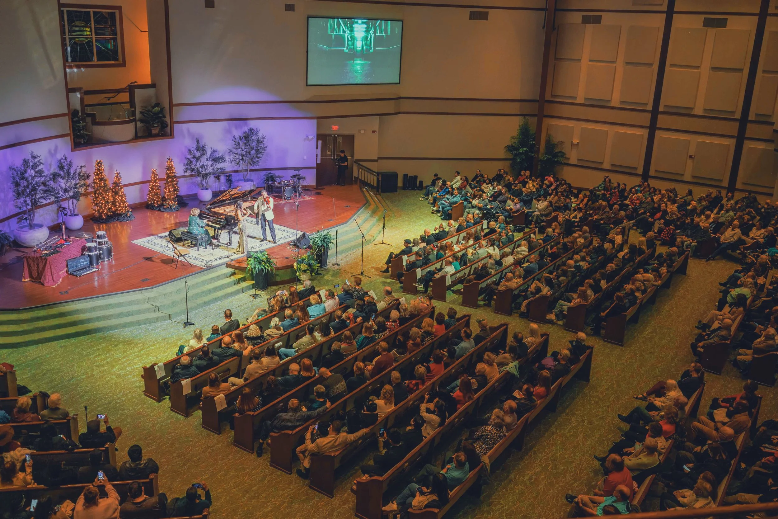 Audience seated in a concert hall watching a performance with a singer and pianist on stage, decorated with Christmas trees and potted plants.